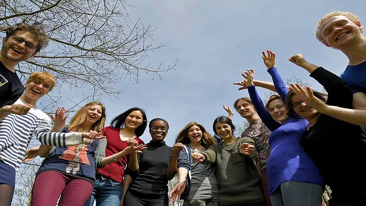 Eine Gruppe junger Studierender stehen im Kreis und winken freundlich in die Kamera.